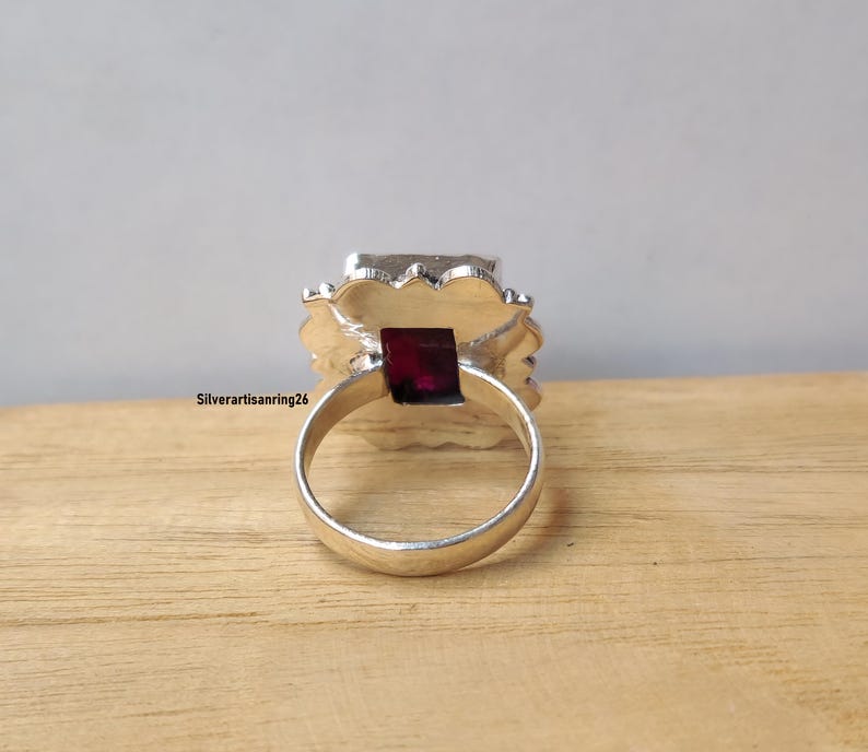 Pu&ograve; includere: Un anello d'argento con una grande gemma quadrata di colore rosso scuro al centro. La gemma &egrave; circondata da un'incastonatura decorativa e strutturata. L'anello &egrave; appoggiato su una superficie di legno. Il testo "Silverartisanring26" &egrave; visibile.