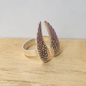 May include: A silver-colored ring featuring an open design with two detailed wing shapes. The wings are positioned to appear as if they are wrapping around a finger. The ring is set against a light wood surface.