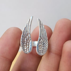 May include: A silver-colored ring featuring a detailed wing design on each side. The wings are open, creating a unique and eye-catching look. The ring is a piece of jewelry.