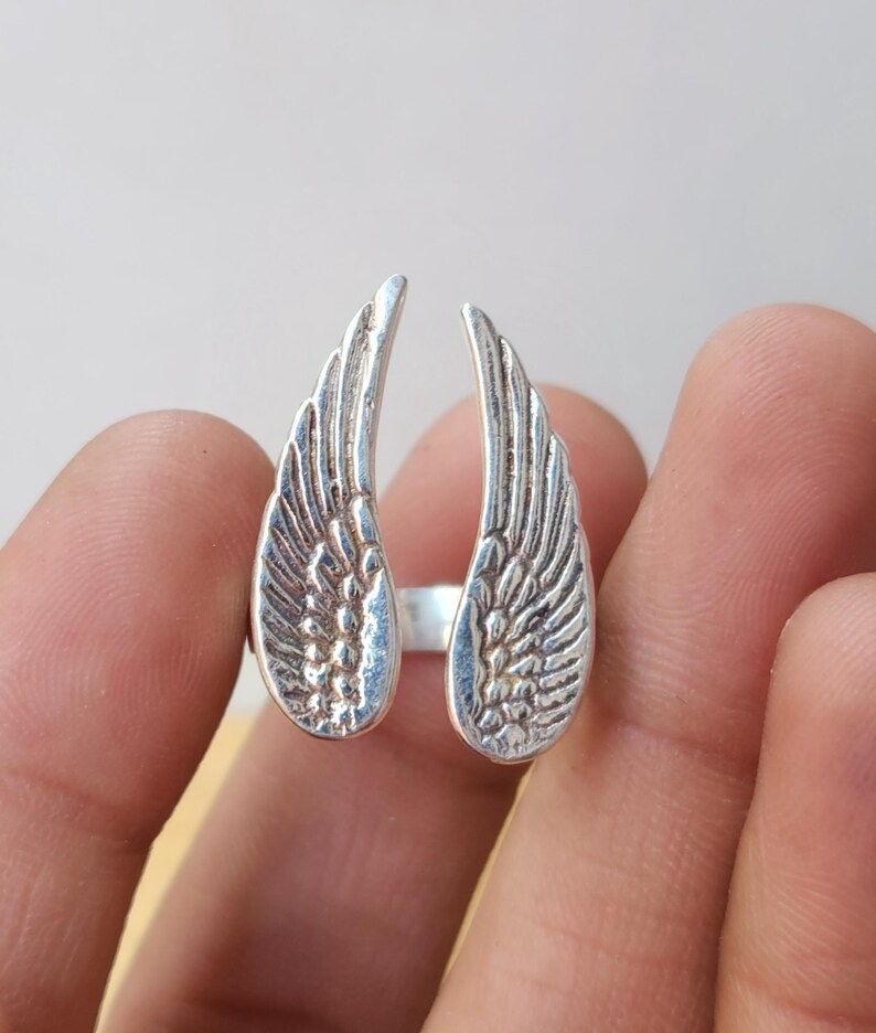 May include: A silver-colored ring featuring a detailed wing design on each side. The wings are open, creating a unique and eye-catching look. The ring is a piece of jewelry.