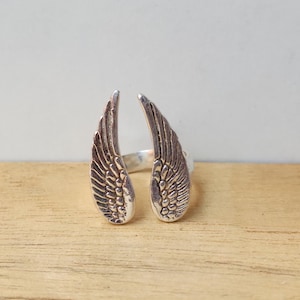 Puede incluir: Un anillo plateado con un diseño abierto, con dos formas de alas detalladas. Las alas están posicionadas para mirar hacia afuera, con intrincados detalles de plumas. El anillo está sobre un fondo claro y una superficie de madera.