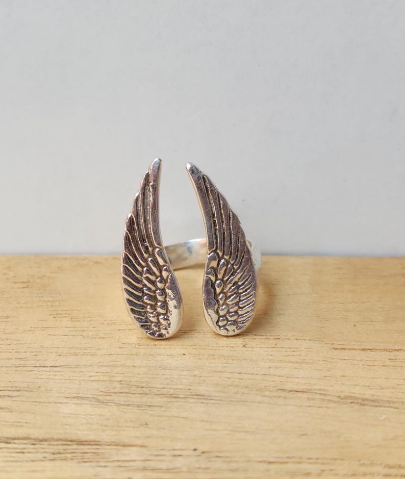 May include: A silver-colored ring featuring an open design with two detailed wing shapes. The wings are positioned to face outward, with intricate feather detailing. The ring is set against a light background and wooden surface.