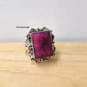 Pu&ograve; includere: Un anello d'argento con una grande gemma rettangolare color magenta. La gemma &egrave; incastonata in una cornice d'argento decorata con un bordo smerlato. L'anello &egrave; esposto su una superficie di legno marrone chiaro.