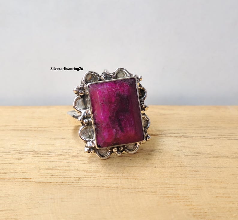Pu&ograve; includere: Un anello d'argento con una grande gemma rettangolare color magenta. La gemma &egrave; incastonata in una cornice d'argento decorata con un bordo smerlato. L'anello &egrave; esposto su una superficie di legno marrone chiaro.