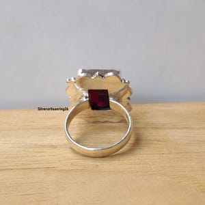 Pu&ograve; includere: Un anello d'argento con una grande gemma quadrata di colore rosso scuro al centro. La gemma &egrave; circondata da un'incastonatura decorativa e strutturata. L'anello &egrave; appoggiato su una superficie di legno. Il testo "Silverartisanring26" &egrave; visibile.