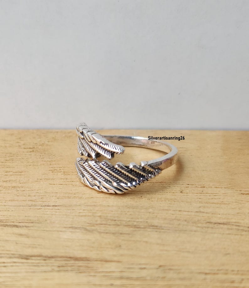 May include: A silver-colored ring with a feather design. The ring wraps around the finger with two detailed feather ends. The ring is displayed on a wooden surface. The text "Silverartisanring26" is visible.