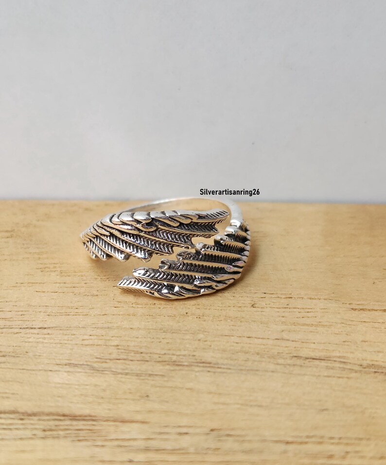 May include: A silver-colored ring with a wing design. The ring features detailed feather-like patterns, with the text "Silverartisanring26" visible. The ring is set against a wooden surface.
