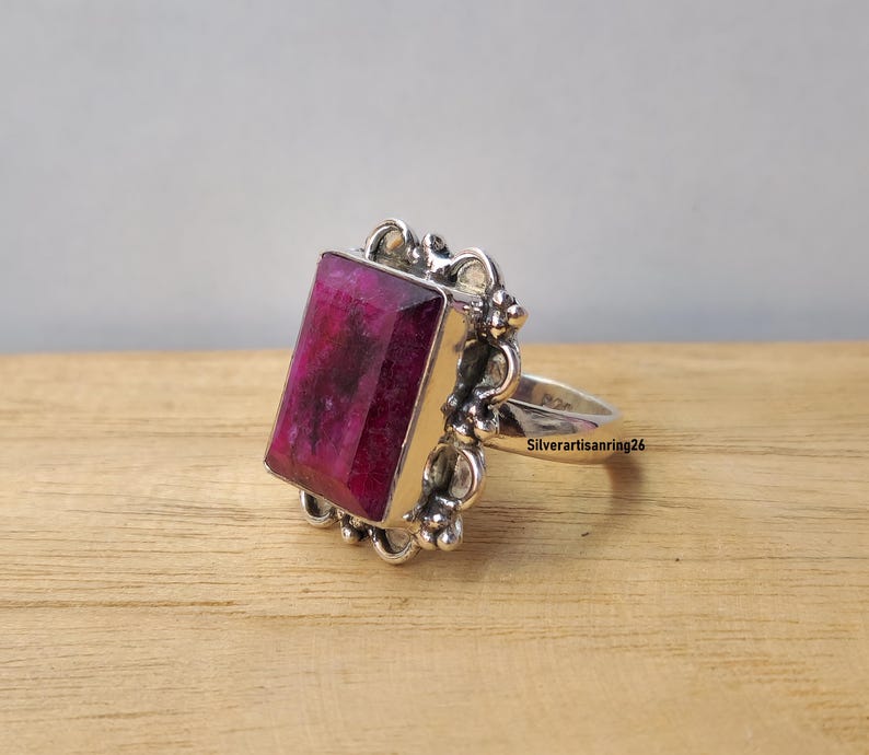 Pu&ograve; includere: Un anello d'argento con una grande gemma rettangolare rosa intenso. La gemma &egrave; incastonata in una montatura d'argento ornata. L'anello &egrave; su una superficie di legno. Il testo "Silverartisanring26" &egrave; visibile sulla fascia dell'anello.