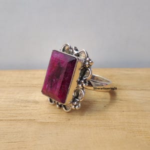 Pu&ograve; includere: Un anello d'argento con una grande gemma rettangolare rosa intenso. La gemma &egrave; incastonata in una montatura d'argento ornata. L'anello &egrave; su una superficie di legno. Il testo "Silverartisanring26" &egrave; visibile sulla fascia dell'anello.