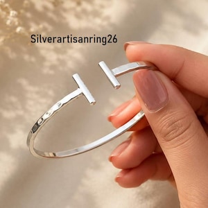 Könnte beinhalten: Ein silberfarbenes, offenes Armband mit einer gehämmerten Textur entlang des Bandes und zwei senkrechten, T-förmigen Stäben an der Öffnung. Der Text Silverartisanring26 ist in der oberen linken Ecke des Bildes zu sehen.