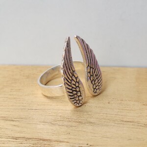 May include: A silver-colored ring featuring an open design with two detailed wing shapes. The wings are positioned to appear as if they are wrapping around a finger. The ring is set against a light wood surface.