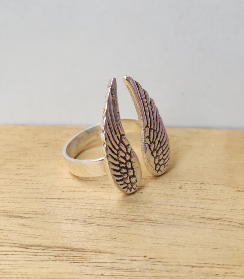 May include: A silver-colored ring featuring an open design with two detailed wing shapes. The wings are positioned to appear as if they are wrapping around a finger. The ring is set against a light wood surface.