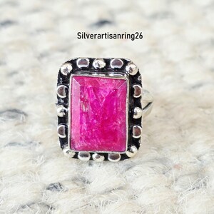 May include: A rectangular ring featuring a vibrant pink gemstone set in a dark, oxidized silver frame with a beaded border. The metal setting displays a textured, darkened finish around the stone. The text Silverartisanring26 is visible.