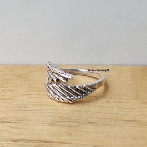 May include: A silver-colored ring with a feather design. The ring wraps around the finger with two detailed feather ends. The ring is set against a light brown wooden surface. The text "Silverartisanring26" is visible.