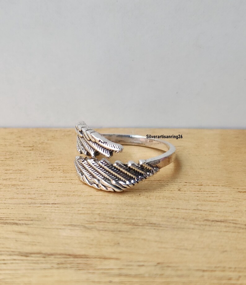 May include: A silver-colored ring with a feather design. The ring wraps around the finger with two detailed feather ends. The ring is set against a light brown wooden surface. The text "Silverartisanring26" is visible.
