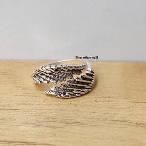 May include: A silver-colored ring with a wing design. The ring features detailed feather-like patterns and is set against a wooden surface. The text "Silverartisanring26" is visible in the background.