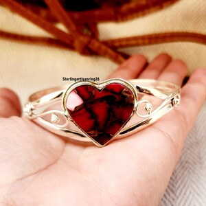 Könnte beinhalten: Ein silberfarbenes Armband mit einem herzförmigen, rot-schwarzen Stein im Mittelpunkt. Das Herz ist von einer silbernen Fassung mit dekorativen Wirbeln umrahmt. Das Armband wird auf einer Hand gezeigt.