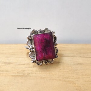 Pu&ograve; includere: Un anello d'argento con una grande gemma rettangolare color magenta. La gemma &egrave; incastonata in una cornice d'argento decorata con un bordo smerlato. L'anello &egrave; esposto su una superficie di legno marrone chiaro.