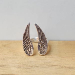 May include: A silver-colored ring featuring an open design with two detailed wing shapes. The wings are positioned to face outward, with intricate feather detailing. The ring is set against a light background and wooden surface.