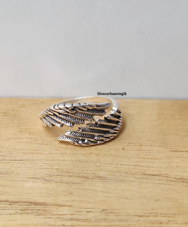 May include: A silver-colored ring with a wing design. The ring features detailed feather-like patterns and is set against a wooden surface. The text "Silverartisanring26" is visible in the background.