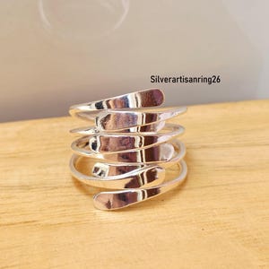 Könnte beinhalten: Ein silberfarbener Metallring mit einem mehrschichtigen Spiraldesign, das aus flachen, überlappenden Bändern besteht, die sich um den Finger wickeln. Die Oberfläche ist poliert und reflektierend. Der Text Silverartisanring26 ist oben rechts sichtbar.