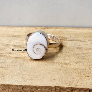 Puede incluir: Un anillo de plata con una piedra preciosa ovalada blanca con un diseño en espiral. El anillo está sobre una superficie de madera. El texto "Silverartisanring26" es visible en la esquina inferior izquierda.