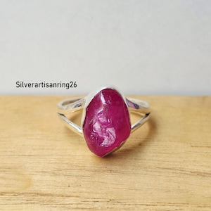 May include: A silver ring featuring a large, raw, deep pink ruby gemstone set in a simple metal bezel. The band of the ring is split into two thin silver wires that meet at the base. The text Silverartisanring26 is visible in the background.