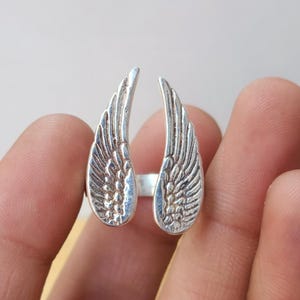 May include: A silver-colored ring featuring a detailed wing design on each side. The wings are open, creating a unique and eye-catching look. The ring is likely made of sterling silver or a similar metal.