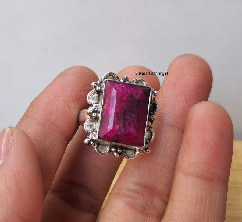Pu&ograve; includere: Un anello d'argento con una grande gemma rettangolare rosa intenso. La gemma &egrave; incastonata in una cornice decorativa smerlata. L'anello &egrave; tenuto in mano, mettendo in risalto gli intricati dettagli del gioiello.