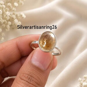 May include: A silver ring featuring a large, oval-shaped, faceted golden-yellow gemstone set in a smooth metal bezel. The band is thin and polished silver. The text Silverartisanring26 is visible in the upper portion of the image.