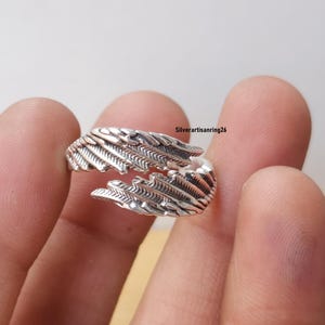 May include: A silver-colored ring with a feather design. The ring wraps around the finger with detailed feather textures. The ring is shown against a neutral background. The text "Silverartisanring26" is visible.