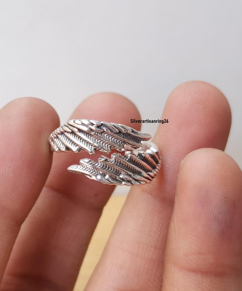 May include: A silver-colored ring with a feather design. The ring wraps around the finger with detailed feather textures. The ring is shown against a neutral background. The text "Silverartisanring26" is visible.