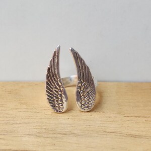 May include: A silver-colored ring featuring an open design with two detailed wing shapes. The wings are positioned to face outward, with intricate feather detailing. The ring is set against a light background and wooden surface.