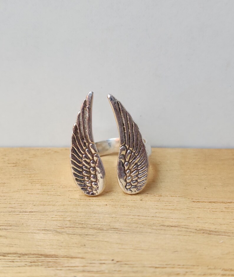 May include: A silver-colored ring featuring an open design with two detailed wing shapes. The wings are positioned to face outward, with intricate feather detailing. The ring is set against a light background and wooden surface.