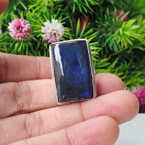 Puede incluir: Un anillo rectangular con una piedra preciosa de labradorita azul oscuro engastada en un marco de metal plateado pulido. La piedra muestra un brillo azul profundo en su superficie. El texto Silverartisanring26 es visible en la esquina superior izquierda.