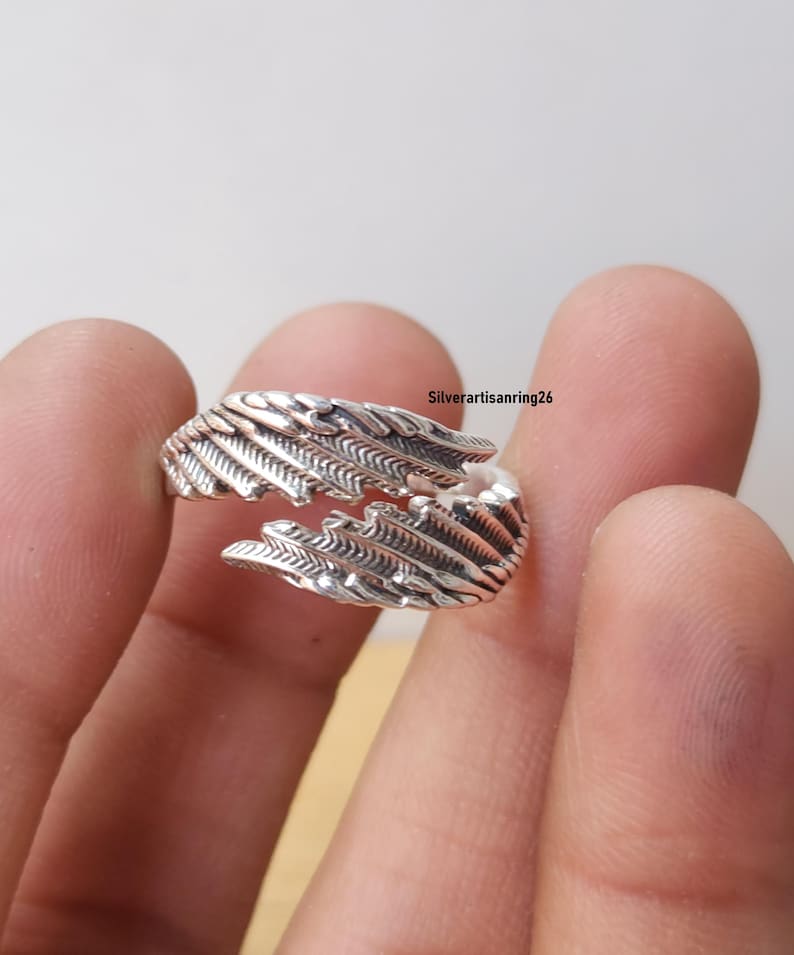 May include: A silver-colored ring with a feather design. The ring wraps around the finger with detailed feather textures. The ring is shown against a neutral background. The text "Silverartisanring26" is visible.