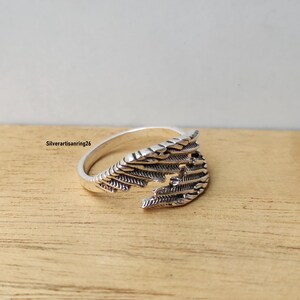 May include: A silver-colored ring with a detailed wing design. The ring features intricate feather-like patterns and is set against a wooden surface. The ring's design is open, allowing for an adjustable fit.