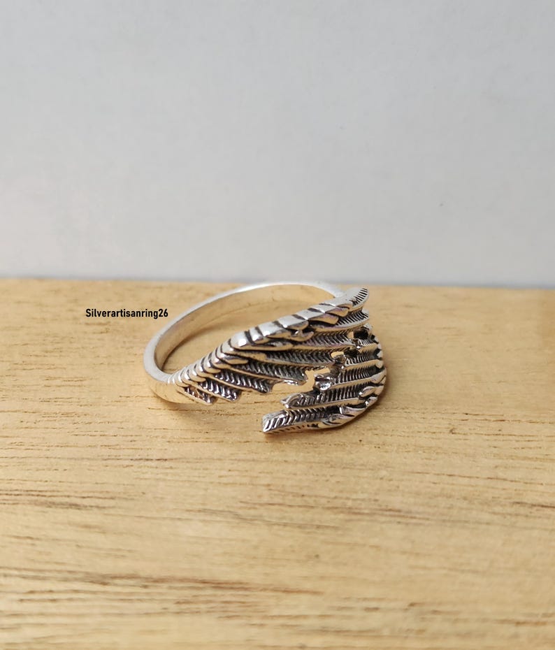 May include: A silver-toned ring with a detailed wing design. The ring features intricate feather-like patterns and is set against a wooden surface. The ring's design is open, allowing for an adjustable fit.