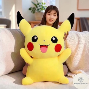 May include: A large yellow plush toy shaped like a cartoon character with long ears tipped in black, round red cheeks, a small black nose, and a wide open mouth showing a pink tongue. The character has two short arms and two short legs.