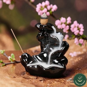 May include: A black ceramic incense burner with a waterfall design. A lit incense cone sits atop the burner, with smoke cascading down the tiers. A lit incense stick is placed in the front. Pink flowers are in the background.