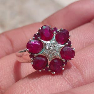 Könnte beinhalten: Ein silberfarbener Ring mit einer Anordnung aus sechs oval geschliffenen, tiefroten Rubinen in einem Blumenmuster um eine sternförmige Fassung, die mit kleinen, klaren, rund geschliffenen weißen Steinen besetzt ist, auf einer polierten Metallschiene.