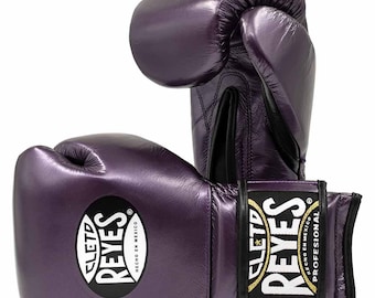 May include: A pair of purple boxing gloves with the brand name "CLETD REYES" in white lettering on a black circle. The gloves are made of shiny material and have a black wrist strap with gold lettering. The gloves are 14oz.