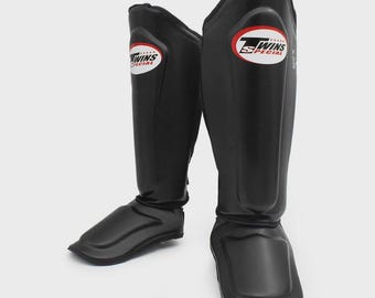 Könnte beinhalten: Paar schwarze Schienbeinschützer mit dem Logo "TWINS SPECIAL" in Weiß und Rot. Diese Schützer sind für Kampfsportarten konzipiert und schützen Schienbeine und Füße. Sie bestehen aus strapazierfähigem Material und eignen sich für Training und Wettkampf.