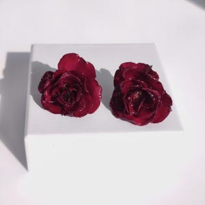 Op de afbeelding: Twee donkerrode roosvormige oorbellen. De oorbellen liggen op een wit oppervlak. De rozen hebben meerdere bloemblaadjes en een glanzende afwerking, met een diepe, rijke kleur. De oorbellen zijn waarschijnlijk voor doorboorde oren.