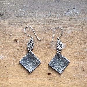 Könnte beinhalten: Ein Paar silberne Ohrringe mit einem gehämmerten rautenförmigen Anhänger. Die Ohrringe haben eine strukturierte Oberfläche und sind an einer geknoteten Kette und einem einfachen Haken befestigt. Die Ohrringe werden auf einer Holzoberfläche präsentiert.