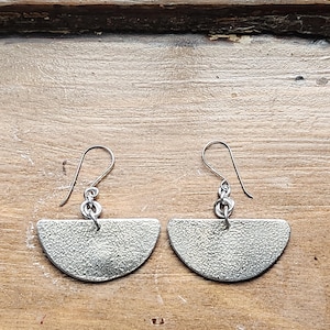 May include: A pair of silver-toned earrings. Each earring features a textured, semi-circular design suspended from a simple hook. The earrings are displayed against a wooden background.