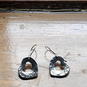 May include: A pair of silver heart-shaped earrings with a hammered texture. The earrings have a simple hook design and a cutout heart shape. The metal has a slightly darkened, aged appearance, and the earrings are displayed on a wooden surface.