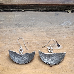 May include: A pair of silver earrings with a hammered texture. Each earring has a curved, crescent-like shape with a hook for pierced ears. The earrings are displayed on a wooden surface.