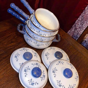May include: A set of white enameled cookware featuring a blue floral pattern. The collection includes three nested pots with blue handles and three matching lids with blue knobs. The interior of the pots is white with a metallic rim.