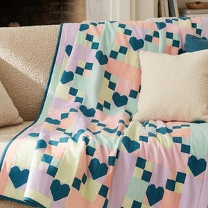 May include: A patchwork quilt featuring a repeating pattern of dark teal hearts interspersed with blocks of light lavender, pale yellow, and soft peach. The quilt is finished with a dark teal border and a grid-like arrangement of small squares.